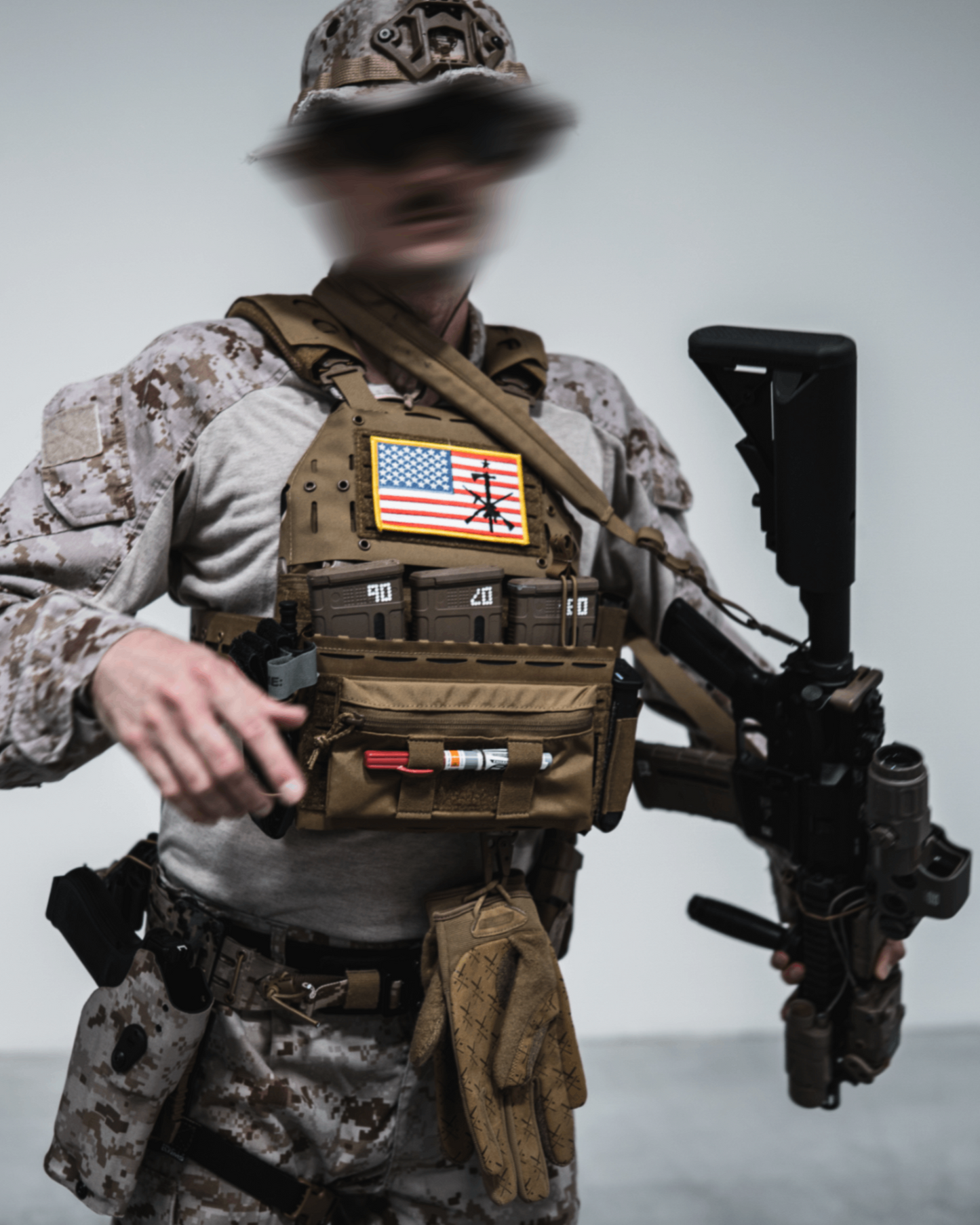 Person in tactical gear with American flag vest and rifles against a plain background